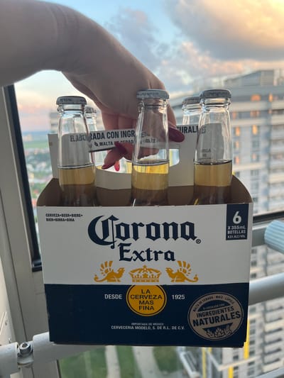 A six-pack of Corona Extra beer bottles is held by a hand with red nail polish against a blurred background of buildings and a cloudy sky at dusk. The bottles are partially obscured by the hand and the cardboard packaging. The packaging displays the Corona Extra logo and information about the product, including the quantity of bottles and alcohol content. The scene appears to be an outdoor setting, possibly a balcony or rooftop, with a city view. The lighting suggests the time is late afternoon or early evening.