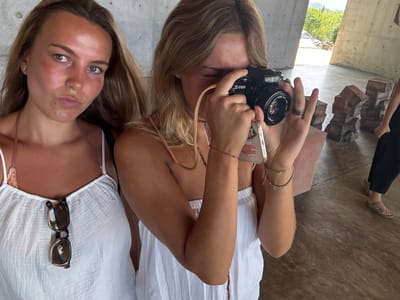 The image captures three individuals within a modern, open-air concrete architectural structure located in San Isidro Llano Grande, Mexico. In the left foreground, a young adult female with light skin and light brown hair looks directly towards the viewer with a slight pout, wearing a white, textured, sleeveless top with a halter strap and sunglasses hanging from her neckline. To her right, a second young adult female, also with light skin and blonde hair, is actively using a black Minolta X-700 film camera, holding it to her right eye, obscuring her face. Her left hand supports a white smartphone with a decorated case behind the camera. She wears multiple rings on her right hand and two chain bracelets and a small red string bracelet on her left wrist. In the background to the right, the lower body of a third individual is visible, wearing dark pants and light-colored sandals, standing near several irregularly stacked, reddish-brown concrete or stone blocks on the light grey concrete floor. The concrete structure features large, smooth grey walls and columns, and an opening in the background reveals a lush green, hilly landscape with trees under a light sky.