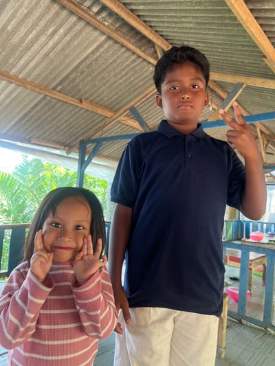 The image depicts two children posing in a semi-open structure located in Gembong, Indonesia. On the right, a boy, approximately 8-12 years old, stands facing forward, wearing a dark blue polo shirt and light-colored shorts. His right hand is raised to shoulder height, forming a "V" sign with his index and middle fingers, while his expression is neutral. To his left and slightly in front, a girl, approximately 4-6 years old, smiles directly at the camera. She wears a pink and white horizontally striped long-sleeved shirt, with both hands playfully positioned against her cheeks, fingers splayed. The overhead structure features a weathered corrugated metal roof supported by visible wooden beams and blue-painted wooden posts. In the background to the left, bright sunlight illuminates green tropical foliage. To the right, a light blue wooden table with a pink bowl and other indistinct items is partially visible.