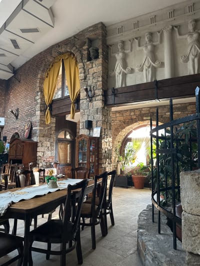 The image depicts an indoor scene, likely a restaurant or a similar establishment, with a rustic and somewhat historical ambiance. The setting is characterized by exposed brick walls, stone accents, and wooden beams. In the foreground, a long wooden table is set with a white tablecloth adorned with a decorative pattern, and several dark wooden chairs surround it. A small potted plant with green leaves sits on the table, along with what appear to be decorative ceramic figures.
The background reveals more architectural details, including arched doorways, a circular window with natural light streaming through, and ornate decorative elements, such as classical-style reliefs on a plastered wall and a decorative iron grate. There are also vintage wooden furniture pieces, including a display cabinet filled with items. The lighting suggests it could be daytime, with natural light illuminating parts of the space. The overall impression is one of a well-decorated, possibly old-world setting, emphasizing texture and natural materials. No people are visible in the image, and there is no clear indication of a specific activity taking place beyond the implied use of the dining area. A small "EXIT" sign is visible near the circular window. The location is identified as Lukovit, Bulgaria.