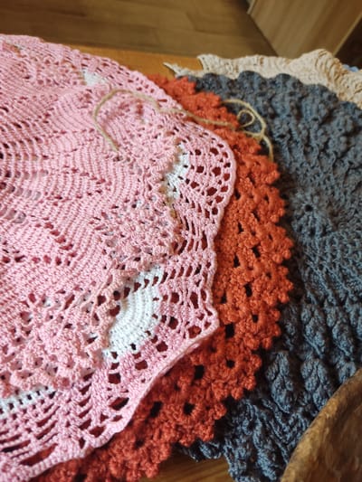 The scene depicts a close-up, elevated view of four distinct crocheted textile items stacked on a light brown wooden surface. The setting for this depiction is the city of Staraya Russa, Russia. Atop the stack is a delicate, light pink crocheted piece featuring intricate open lacework and integrated white sections, exhibiting a scalloped or wavy edge. A piece of natural light brown twine rests on this pink textile. Directly beneath the pink item is an orange-rust colored crocheted piece with a dense, textured pattern, possibly a series of repeating floral or square motifs. Further down the stack, a dark grey or deep blue crocheted textile is partially visible, characterized by a highly textured surface of raised stitches, potentially bobbles or popcorns. The final, topmost visible item in the stack is a beige or cream-colored crocheted piece, partially obscured and displaying a more open, looser pattern. The immediate background shows a blurred wooden surface, and a curved, dark brown wooden object, possibly a bowl, is partially visible and out of focus in the bottom right corner.