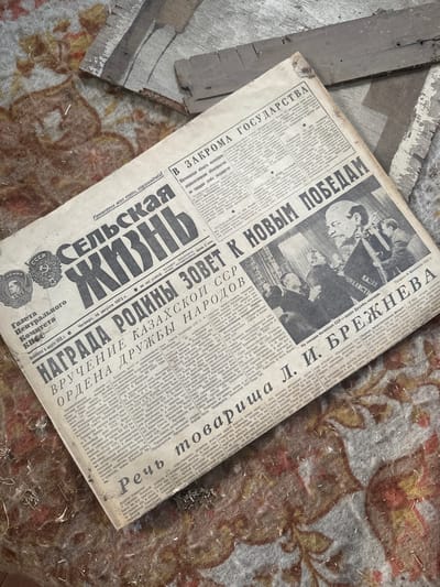The image shows a discarded newspaper titled "СЕЛЬСКАЯ ЖИЗНЬ" (Rural Life) in Russian, dated August 16, 1973. The newspaper features an image of Leonid Brezhnev and articles about achievements in agriculture and national awards. The location appears to be in a state of decay, with the newspaper resting on a damaged, floral-patterned surface and pieces of broken wood nearby. The photograph was most likely taken in Студзёнкаўскі сельскі Савет, Belarus.