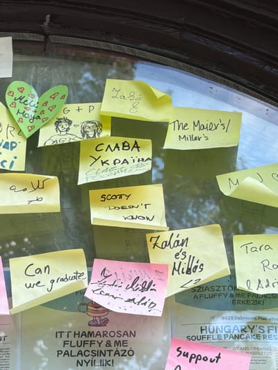 This is a medium shot photograph, taken at eye level, of a window pane covered with numerous handwritten sticky notes and some drawings. The window pane appears to be part of an establishment's display or notice board. The notes are of various colors, including yellow, green, pink, and white, and are written in different languages, such as English, Ukrainian, and Hungarian, as well as Arabic script. Some notes contain drawings, including a heart, two people, and a cartoon character holding a mug. One yellow sticky note has "Meli & Maya" written inside a green heart. Below that is a drawing of two people with hearts between them and "G+P". Another yellow note says "СЛАВА УКРАЇНІ" and below it "СЛАВА РОССИИ". A yellow note in Arabic script reads "كلامه". Another yellow note says "SCOTY DOESN'T KNOW". A pink note has some indecipherable text. A yellow note has "Zolán e's Milós". Another yellow note has "Tara Ro <3 Adri". In the background, a poster is visible with text in Hungarian, including "ITT HAMAROSAN FLUFFY & ME PALACSINTÁZÓ NYÍLIK!" and "HUNGARY'S FI SOUFFLE PANCAKE RES". A small logo with an address is visible on the poster: "4025 Debrecen, Piac utca". The overall environment appears to be outdoors or in a semi-outdoor setting, with greenery visible through the window and soft, diffused lighting. The image is slightly out of focus, particularly in the background.