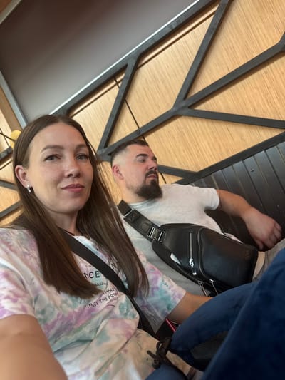 The image captures two individuals, seemingly in a casual setting. A woman with long brown hair is taking a selfie, with a man lying behind her. The man has a beard and is wearing a white t-shirt and a black crossbody bag. They are positioned near a structure with a geometric pattern made of wood panels and dark metal framing. Part of a blue object is also visible in the background. The clothing and accessories suggest a contemporary style. Based on the text on the woman's shirt, they are located in Bol'shie Kabany, Russia, specifically at coordinates E 49.62 km.