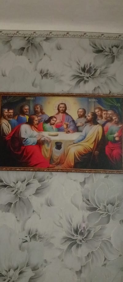 This is a framed painting depicting "The Last Supper." The central figure, Jesus, is seated at a table with twelve disciples. Jesus, distinguished by a halo and dressed in a red and purple robe, holds a golden chalice. The disciples, in various colored robes, are seated around him, some appearing to be in conversation or contemplation. One disciple, to Jesus' left, rests his head on his hand, conveying a sense of sadness or weariness. On the table, there are loaves of bread and a dark goblet. The scene is set indoors, with a dark background that is softly illuminated by a golden light emanating from behind Jesus. The painting is framed by a simple, ornate gold border. The painting hangs on a wall covered in a floral wallpaper. The wallpaper features a repeating pattern of large, stylized flowers in shades of grey and white, with subtle darker shading to create depth and dimension. The wallpaper's design is delicate and abstract, contrasting with the detailed religious scene of the painting. The image appears to be taken in natural indoor lighting. There is no visible text in the image.