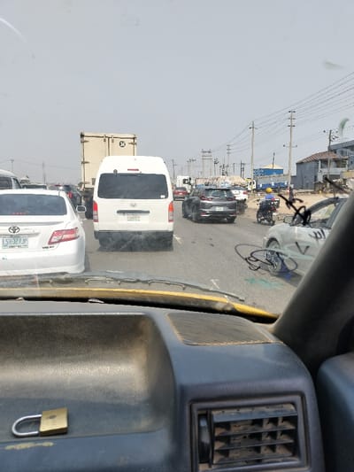 The image depicts a congested multi-lane road in the city of Magado, Nigeria, viewed from inside a vehicle through its windshield. The sky is hazy and bright.
In the immediate foreground, the dark-colored dashboard of the camera vehicle is visible, featuring an air vent on the right and a gold-colored padlock resting on its surface. The bottom edge of the windshield and the top of the wipers show signs of wear and dirt.
Ahead, in the left lane, a white Toyota Camry sedan (license plate LSR-327JP) is directly in front. To its right, in the center lane, a white Toyota Hiace van (license plate HJ685FH) precedes it. Further right, a light-colored sedan is partially visible, displaying dark, linear markings on its windshield. A black motorcycle with a rider wearing a yellow helmet is observed to the right of the van, navigating through traffic. A dark gray SUV (Honda CRV) is also visible further ahead in the lane to the right of the van. Numerous other vehicles, including a beige cargo truck marked "34" on its rear, are stationary or moving slowly in the distant lanes.
The environment features utility poles carrying multiple power lines running parallel to the road on both sides, extending into the background. Buildings, including one with a blue shipping container on its roof, line the roadside. The overall scene suggests heavy urban or semi-urban traffic conditions under a hazy sky.