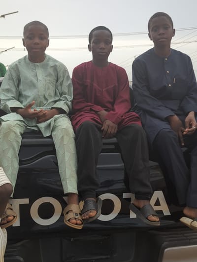 Three young men are sitting on the tailgate of a black Toyota pickup truck. The tailgate is marked with "TOYOTA" in large white letters. The truck is parked in an outdoor environment, likely in Kafanchan A, Nigeria. The men are sitting in a row from the front of the truck, and the camera is at a slight angle. The man in the front is wearing a light-green patterned robe and matching pants. The man in the middle is wearing a maroon-colored robe and black pants. The man in the back is wearing a dark blue robe and matching pants. All three men are wearing sandals.