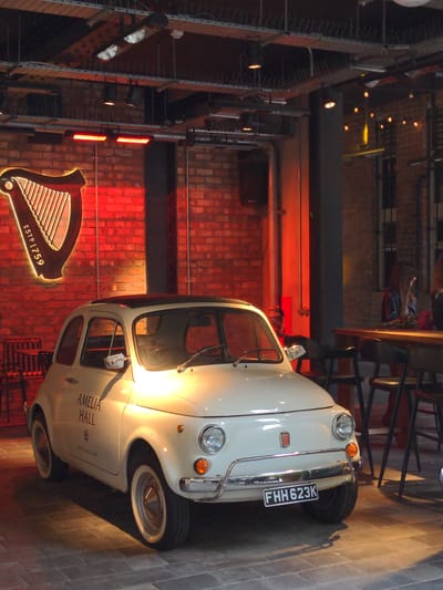 The image depicts the exterior of a white Fiat 500 parked on a brick-patterned floor. The car has its headlights on and a license plate that reads "FHH 623K". To the left of the car is a large, illuminated Guinness harp logo against a brick wall. The background reveals an indoor setting with industrial-style lighting fixtures on the ceiling and exposed brick walls. In the far right, a table with chairs is visible, where two women are seated. One woman, with dark hair, is wearing a black leather jacket and looking towards the table. The environment suggests a bar or restaurant, likely in Belfast, United Kingdom, given the prominent Guinness branding.