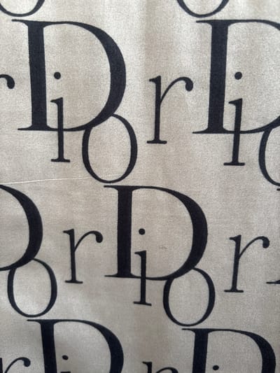 The image is a close-up photograph of a beige fabric with a repeating pattern of large, dark blue letters that appear to spell out "Dior". The pattern is dense, with letters overlapping and arranged in a way that creates a textured effect. The fabric itself has a slight sheen, and the weave of the material is visible. There are no people or any recognizable environmental elements. The image does not provide any information about its location, including in the city of Imeni Morozova, Russia.
