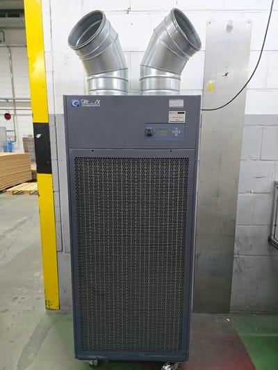 The image shows a large, dark gray industrial dehumidifier with two silver cylindrical ducts on top. The dehumidifier has a perforated front panel and a control panel with buttons and a small screen. It is located indoors in what appears to be a warehouse or industrial setting, with a concrete floor and a white brick wall in the background. A yellow and black striped post is visible to the left of the dehumidifier. The scene does not contain any people or visible actions. The location is not specified as being in Worms, Germany.