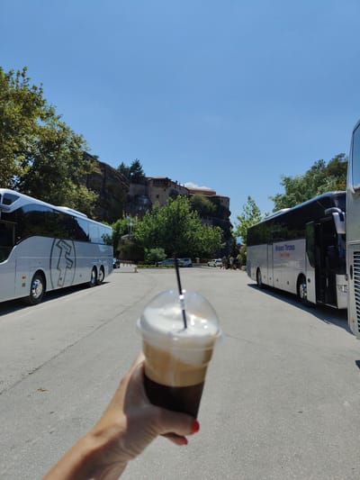 A hand holds a Frappuccino in a clear plastic cup with a straw and white foam topping, in the foreground, left of center. The cup is out of focus. The background reveals a paved parking lot with two tour buses parked. The bus on the left is silver with a black stripe and a circular logo with the letters "T. TOURS" in a stylized font. The bus on the right is silver with black windows and text that reads "Meteor Thorne Travel Center." Behind the buses, a rocky cliff face rises, topped with ancient stone buildings, likely monasteries, which are characteristic of Meteora in Kalambaka, Greece. Green trees partially obscure the rock formations and buildings. The sky is clear and blue.
