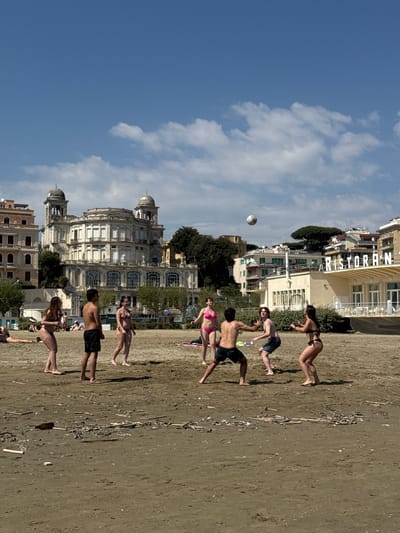 A group of people is playing with a soccer ball on a beach in Anzio, Italy. The people are wearing swimsuits. The beach is sandy and there are some scattered pieces of debris. In the background, there are buildings, including a restaurant. The sky is blue with some clouds. The photograph is taken at an angle, with the buildings appearing to be tilted.