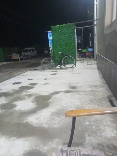 The image depicts an outdoor scene at night, likely in Shar-Bakr, Uzbekistan, with a concrete ground surface that appears wet in patches. In the foreground, the armrest and part of the seat of a wooden chair are visible, with a patterned fabric upholstery on the seat. To the left, a bicycle with black frame and silver wheels is parked in front of a tall, green, hedge-like decorative panel. Behind the bicycle and panel, a structure with metal bars is visible, supporting what appears to be a light fixture. Further back, a white van is parked. To the right of the van, a smaller dark-colored vehicle is also present. In the far background, there are indications of buildings and trees. The overall impression is of a somewhat unkempt or service area at night.