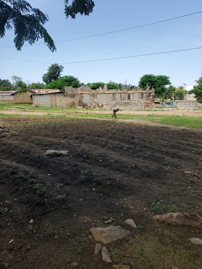 The image captures an outdoor scene in Kabwir, Nigeria. In the foreground, a patch of dark, dry earth is visible, with scattered green vegetation and a few rocks. To the left, a partially constructed building is in progress, with concrete blocks and wooden scaffolding evident. A person is seen walking near the construction site. Beyond the building, trees and more constructed elements can be observed, including corrugated metal roofing. The sky is a clear blue, suggesting it is daytime with fair weather. The overall impression is one of development and ongoing construction in a rural or semi-rural setting.