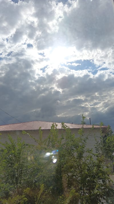The image shows a view of a building with a red roof, partially obscured by green foliage in the foreground. The building appears to be constructed from white brick. Above the building, the sky is filled with a mix of clouds and patches of blue. The sun is visible, shining brightly through some of the clouds, creating a lens flare effect on the foliage in the lower part of the image. The lighting suggests it is daytime, likely late morning or early afternoon, with a partly cloudy and possibly bright weather condition. The foliage suggests an outdoor setting, possibly a residential or semi-rural area. There is no visible text in the image.