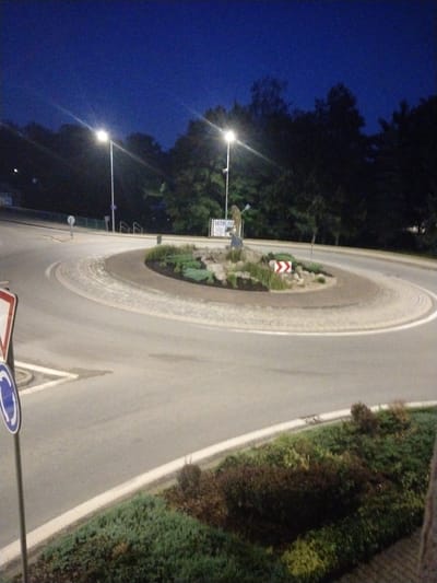The image shows a roundabout at dusk in Častolovice, Czechia. A white street lamp illuminates the scene, casting long shadows. The roundabout itself is paved with cobblestones and features a small garden in the center, complete with rocks, plants, and a statue of a figure holding what appears to be a blue flag or scarf. A red and white chevron sign, typically indicating a hazard or obstruction, is placed on the roundabout's garden island. A circular blue and white traffic sign, denoting a roundabout, is visible on a pole to the left of the image. The surrounding area consists of trees and some low-lying bushes. The sky is a deep, dark blue, indicative of twilight. No people are visible in the image.