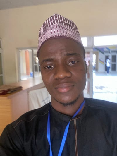 An adult male, appearing to be of West African descent, is depicted in a medium-close-up, selfie-style photograph, taken within a building in Baganakwo, Nigeria. The male subject occupies the central and lower portions of the frame, directly facing the camera with a neutral expression.
He wears a dark, possibly black or deep navy, collared tunic with visible light-brown or orange stitching along the placket and shoulder seams. A royal blue lanyard is draped around his neck. On his head is a maroon and white kufi or prayer cap, featuring an intricate geometric woven pattern. He has dark skin, dark brown eyes, and a neatly trimmed dark beard and mustache.
The background suggests an indoor or sheltered transitional space, possibly a hallway or open-plan room with light beige or off-white walls. To the subject's left, a light-colored, possibly wooden, low partition or counter is visible, on which a folded, patterned textile (predominantly red and brown) is placed. Beyond this, an open doorway or archway leads to another room or corridor, revealing light-colored walls and a vertical brown architectural element. A blue object is faintly discernible in the distant left background. To the subject's right, the background continues with similar light-colored architectural elements. Further into the distance, a blurry figure of an individual in a white top and dark lower garment is partially visible, appearing to walk or stand near a doorway or structure. Faint, out-of-focus colorful signage or banners are visible on the far wall. The scene is brightly and evenly lit, consistent with ambient daylight.