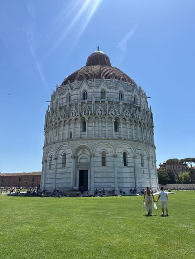 The image depicts the Pisa Baptistery in Pisa, Italy, a circular marble building with a domed roof. It is situated on a vast, manicured green lawn where numerous people are spread out, some sitting and others lying down. A couple, a man and a woman, are walking hand-in-hand away from the viewer towards the Baptistery. The man is wearing a white polo shirt and white shorts, and the woman is wearing a beige outfit. They are holding bags. The sky is a clear, bright blue with a prominent sun flare in the upper left quadrant. In the background, other buildings and trees are visible.