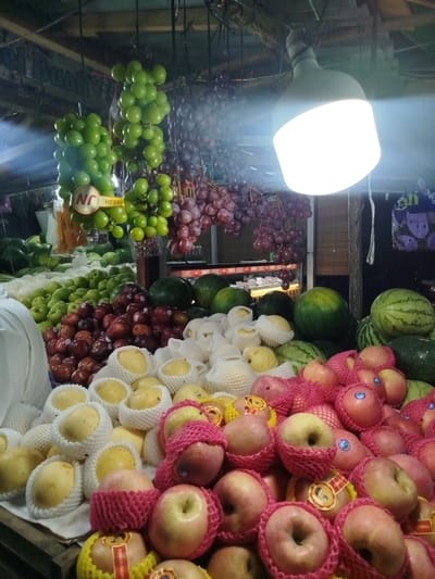 The image depicts a fruit stall, likely in the city of Kutal, Sukon, Indonesia, illuminated by an overhead lamp. The stall displays a variety of fresh fruits, including bunches of green and red grapes hanging from above. Piles of watermelons and green apples are visible in the background, while the foreground features neatly arranged pears individually wrapped in protective foam netting and several red apples also covered in pink netting. Some fruits have stickers indicating they are for sale. The setting appears to be an outdoor market, with a rustic wooden structure overhead and a glimpse of a display cabinet in the background. The overall impression is of a vibrant and well-stocked fruit vendor.