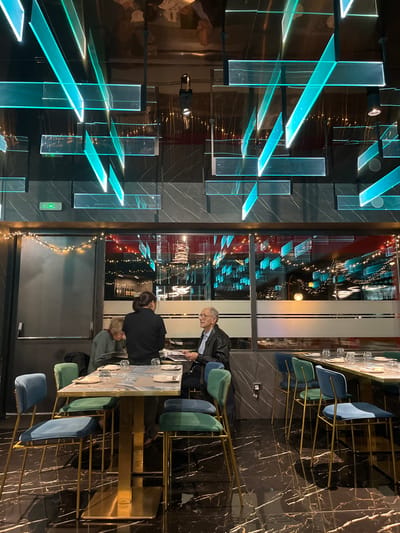 This media file captures an elegant, modern restaurant or bar interior, likely in the evening given the ambient lighting and dark reflections outside.

The scene focuses on a dining area with a long, rectangular table featuring a striking gold-colored base. Around this table are several upholstered chairs in shades of teal, blue, and green, some of which are partially obscured. Three individuals are visible at the table: an older man with glasses, dressed in a dark jacket over a lighter shirt, stands at one end, looking down at a menu or document on the table. Two other people are seated; one is seen from behind with dark hair tied back, and another with lighter hair is partially visible in profile. Glassware is set on the table, indicating a dining or socializing activity.

A prominent design feature dominates the left side of the frame: an intricate installation of numerous translucent glass panels, dramatically illuminated with a vibrant, cool blue-green light. These panels are arranged geometrically, casting an ethereal glow and creating striking visual patterns against the dark, possibly marbled, wall.

A large window spans across the image, reflecting the interior's illuminated glass art and offering a glimpse of indistinct distant lights outside. String lights are draped near this window, adding a subtle warmth to the cool-toned decor. The overall aesthetic is sophisticated and contemporary, characterized by rich textures, metallic accents, and artistic lighting. No discernible text is visible in the image.