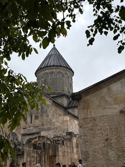 An exterior, medium shot captures a stone church structure with a conical roof visible through the branches of a tree. The church is constructed of rough-hewn stone blocks in shades of beige and gray. The main section of the building has a domed roof topped with a spire, and intricate stone carvings adorn its facade. To the right of the main structure, a wall of lighter-colored stone extends upwards, with a dark gutter running along its top edge. In the foreground, the lower part of the church features arched entrances and a section of ruined stonework, partially obscured by foliage. Several individuals are present in the lower portion of the frame, observing the structure. The sky above is overcast and gray.