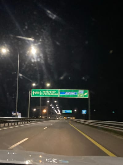 The image shows a view from a vehicle driving on a highway at night. There are overhead highway signs. The sign on the left indicates an upcoming exit to "C. ПЕТЕРБУРГ ST. PETERSBURG" with an arrow pointing up and the number "R10". To the right of this sign, another sign indicates "РОССИЯ" (Russia) with the number "M10" and "E105" written below. The highway is illuminated by streetlights and the headlights of other vehicles. The scene appears to be on a multi-lane highway with concrete barriers and guardrails. The weather appears clear.