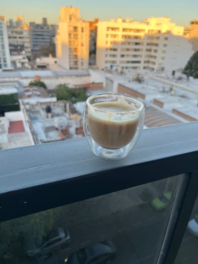 A coffee drink in a glass is on a ledge overlooking a cityscape, likely in Cordoba, Argentina. The drink is in a double-walled glass, with a visible layer of foam or milk on top of the coffee. The ledge appears to be a balcony railing, made of metal. The background consists of buildings, mostly white or light-colored, characteristic of Cordoba architecture. The buildings vary in height, with some taller structures in the distance. The light suggests either early morning or late afternoon, casting a warm glow on some of the buildings. The overall scene conveys a sense of urban tranquility and a moment of relaxation.