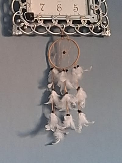 The image displays two decorative objects mounted on a light blue-grey wall. In the upper portion, the visible top section of a rectangular clock is present, featuring an ornate silver-colored metallic frame with intricate scrollwork. Its white face partially displays the Arabic numerals "7," "6," and "5." Directly below this, a dreamcatcher hangs. It consists of a circular, light-brown hoop containing a single dark bead in its center. Multiple strands descend from the hoop, each terminating in a cluster of white feathers, typically accented with a small dark bead or cap at the feather cluster's origin. Shadows are visible on the wall to the right of the objects, indicating a light source from the left. No individuals are depicted. The specific location within Selyanin, Bulgaria, is not discernible from the visual information provided.