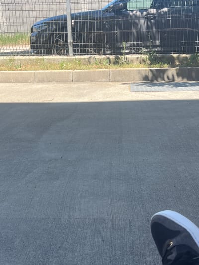 In Tsaratsovo, Bulgaria, a black car is parked behind a wire fence. The car's front end and a portion of its side are visible. The fence is made of vertical metal bars. In front of the fence is a concrete curb with grass and weeds growing along its edge. Beyond the curb is a paved area, likely a sidewalk or road, with distinct concrete lines. A shadow from an overhead object falls across this paved surface. In the lower right corner of the image, a black and white shoe is visible, suggesting a person is present but not fully depicted. The overall scene appears to be an outdoor urban or suburban environment.