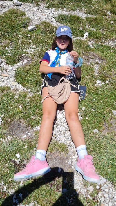 A young girl is lying on her back in a grassy, rocky area, likely outdoors in a natural setting. She is wearing a blue baseball cap, a scout-like neckerchief with blue, yellow, and white patterns, a t-shirt with blue and red sleeves and a white front, and black shorts with white trim. Her feet are adorned with white socks with blue stripes and bright pink sneakers with light blue soles and accents. She is holding a clear plastic water bottle in her right hand. A small, light brown fanny pack is visible around her waist.

The lighting suggests it is daytime, possibly late morning or early afternoon, with the sun casting a prominent shadow to the right of the frame. The weather appears to be clear and sunny, conducive to outdoor activities. The ground is covered with a mix of green grass, small wildflowers, and scattered grey and white rocks, typical of a mountain or hillside terrain.

The girl's expression is relaxed, with her mouth slightly open, and her eyes are looking upwards. She appears to be resting or taking a break. The overall impression is one of a young person enjoying an outdoor adventure, possibly hiking or camping. No discernible text is visible on the girl's clothing or accessories, other than a possible logo on the blue cap. The provided location context of L'Aquila, Italy, further supports the interpretation of an outdoor, mountainous environment.
