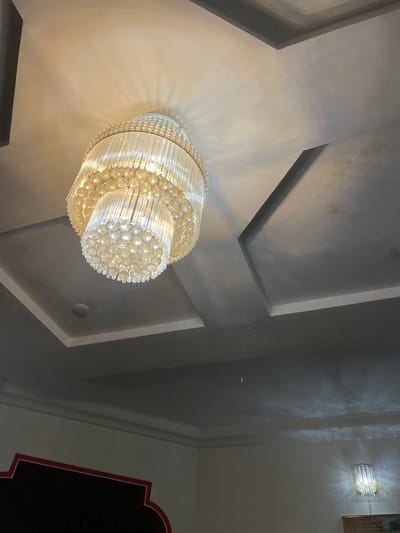 A low-angle indoor shot captures a luxurious chandelier made of glass rods and circular crystal beads, suspended from a ceiling. The ceiling features recessed panels with grey trim. To the right, a wall sconce with similar crystal detailing is illuminated. The lower portion of the image shows a dark wall with a decorative red and white border. The location is Baccure Kofare, Nigeria.