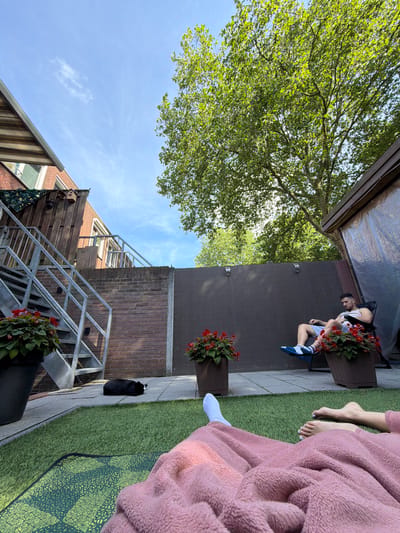 A low-angle shot shows a bright, sunny day with a clear blue sky and dappled sunlight filtering through lush green tree leaves. In the foreground, a person's legs are covered by a soft, pink fuzzy blanket, with one white sock visible. The person's bare feet are peeking out, with light blue nail polish on their toes. A black cat is lying on a grey concrete pathway next to a bright green artificial lawn.
Behind the cat, a dark grey brick wall rises, with metal outdoor stairs leading up to a wooden balcony. A black birdhouse is attached to the wooden fence on the balcony. Red flowers in wooden planters add splashes of color to the scene.
Further to the left, a man is seated on a wooden bench, looking down. He is wearing a white t-shirt, blue shorts, and white and blue sneakers. Red flowers are also planted in a wooden planter next to him. The background shows a brown textured wall and a clear blue sky with a few wispy clouds. The overall atmosphere suggests a relaxed, outdoor gathering on a warm day. The image is oriented such that the sky appears at the bottom, and the ground at the top, indicating it was taken with the camera rotated. The location is Helmond, Netherlands. There is no visible text in the image.