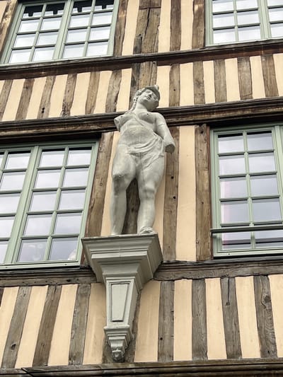 A weathered half-timbered building facade, characteristic of structures in Rouen, France, is depicted from a low angle. The facade consists of dark, irregular vertical and horizontal wooden beams framing light beige or cream-colored infill panels. Three multi-paned windows are visible: two on the upper level and one below them, offset to the left. Each window features light sage green frames dividing the glass into twelve rectangular panes, which reflect an overcast sky.

Centrally positioned on the facade, beneath the upper windows and above the line of the lower window, is a light-grey stone statue of a female figure. The statue is damaged, with both arms missing from below the shoulders. Its head is tilted slightly upward and to the viewer's right. The figure is partially draped in fabric around its lower body, leaving the upper torso partially exposed. The statue rests on a decorative, light-grey corbel that projects from the building. A prominent dark horizontal timber beam separates the upper and lower sections of the facade, running above the statue's corbel. The wooden timbers show signs of age, including cracks, knots, and variations in texture.