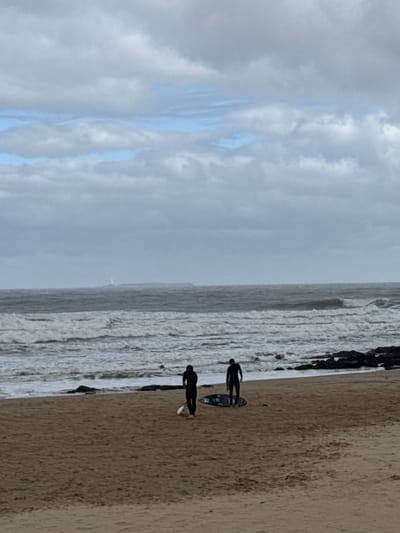 The image displays a beach scene in Punta del Este, Uruguay, characterized by an overcast sky. Two individuals, silhouetted in dark wetsuits, stand on the sandy shore near the waterline. One person is positioned upright, holding a dark surfboard vertically, while the other is stooped or kneeling beside a second surfboard, which has a lighter underside.
To the right of the sandy beach, the ocean stretches out, exhibiting active, choppy waves with visible whitecaps. A dark line of what appears to be rocks or a groyne extends into the water behind the two figures. The sky, filling the upper portion of the frame, is largely cloudy, though some streaks of lighter blue are visible, suggesting breaks in the cloud cover. The diffused light implies either an early morning, late afternoon, or a heavily overcast day.
The primary activity depicted is the presence of surfers observing the ocean, potentially preparing to enter the water or having just finished a session. No specific emotions are discernible from their distant, silhouetted forms. There is no visible text in the image.