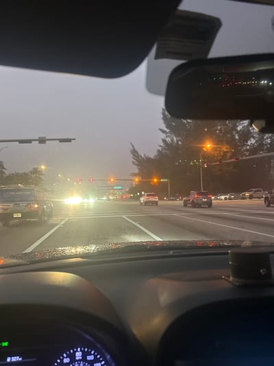 The image presents a wet, overcast street intersection at NW 97th Ave and NW 20th Street in Doral, Florida, United States, viewed from the driver's seat of a stationary vehicle. Raindrops are visible on the vehicle's dashboard and windshield, indicating recent precipitation. The foreground shows the top of the dashboard, a portion of the steering wheel and instrument cluster displaying "327mi," a lowered sun visor on the left, and a rearview mirror on the right reflecting distant lights. A dark cylindrical object is affixed to the right side of the dashboard.
In the midground, multiple vehicles are stopped at the intersection, illuminated by their headlights and streetlights. Traffic lights on the left display red, while those on the right are amber. A light-colored SUV with "FREE INSPECTIONS" written on its rear is visible in the left lane. Overhead street signs confirm the intersection as "NW 20 ST" and "NW 97 AVE," with a more distant green sign partially obscured by atmospheric haze. The background features palm trees on the left and dense dark foliage on the right. No individuals are discernible within the frame. The overall scene suggests an urban setting during evening or early morning hours with low visibility conditions due to the weather.