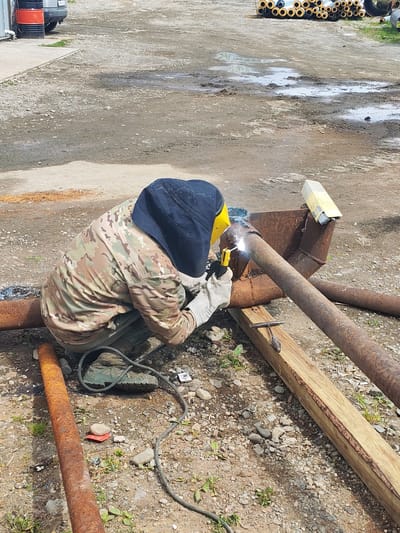 In Termal'nyy, Russia, a person wearing a camouflage jacket, green pants, thick gloves, and a welding mask with a yellow shield is kneeling on the ground. They are actively welding a rusty metal pipe with a welding torch, producing a bright spark. The pipe is supported by a wooden beam. Scattered around are other rusty pipes, gravel, dirt, and some puddles of water. In the background, a pile of insulated pipes and a vehicle can be seen. The scene appears to be an outdoor work area.