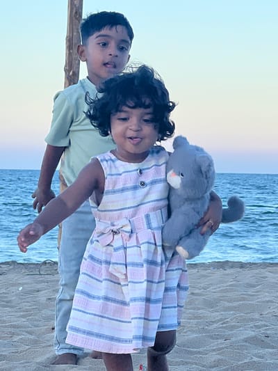 Two children stand on a sandy beach next to calm water. The younger child, wearing a striped dress, carries a gray stuffed animal. The older child, to the left, wears a light green shirt. The location is Kalmunai, Sri Lanka.