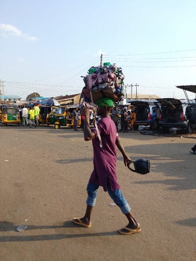 A man carries a large bundle of items on his head, walking across a paved area in what appears to be a bustling marketplace. He wears a green cap, a long purple shirt, and cuffed jeans, along with sandals. In his hand, he carries a black container with a handle. The background features auto-rickshaws, cars with open trunks, and people milling about, suggesting a busy scene. Overhead wires and light-colored buildings are also visible.