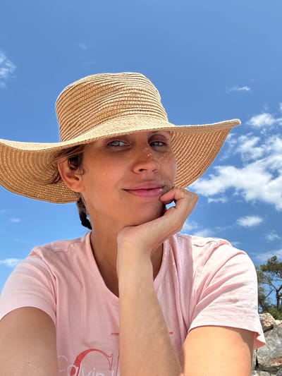 A woman is photographed from a low angle, gazing directly at the camera. She is wearing a wide-brimmed straw hat, casting shadows on her face. She has a pink t-shirt on. Her hand is near her face, seemingly touching her cheek. The backdrop is a bright blue sky with scattered white clouds. Based on the presence of characteristic trees and rocks, this image could have been taken in the city of Ortaca, Turkey.