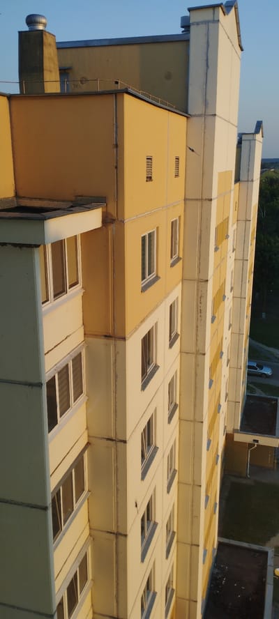 The image displays a close-up, high-angle view of a light yellow and cream-colored apartment building with multiple stories. The building has numerous windows, most of which are rectangular and have light-colored frames. The facade shows signs of age with visible joint lines and some discoloration. On the left side of the building, there is a balcony with windows. At the top of the building, there are rooftop structures, including a chimney with a metallic cap. The building is situated in an urban environment with a clear blue sky. In the background, there are trees and a road with a silver car driving on it. The overall scene appears to be a residential area in Skidzieĺ, Belarus.