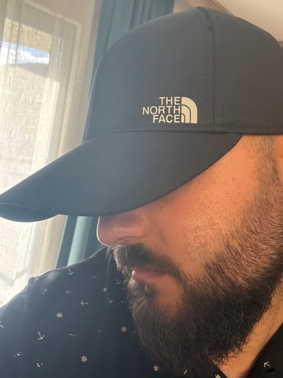 A close-up, high-angle, medium shot shows the side profile of a person wearing a black baseball cap with "THE NORTH FACE" logo. The cap is pulled down, obscuring the upper half of the face, revealing only the nose, mouth, and a full beard. The person is wearing a dark blue shirt with a pattern of white anchors and ship wheels. The background consists of a window with sheer curtains and a glimpse of the outdoors. The lighting suggests the scene is indoors, possibly in Ayvalık, Turkey, given the context.