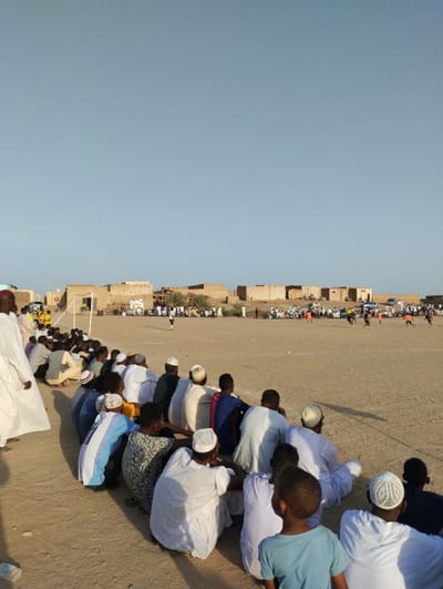 A soccer game unfolds on a barren field under a vast, clear sky. A crowd of spectators, mostly men in traditional white garments and head coverings, sit on the ground, watching the action. Buildings line the horizon, suggesting a rural or desert community. The field is dry and dusty. Players can be seen in uniform, moving with the ball, while more onlookers gather behind the goalposts. A yellow car is parked nearby.