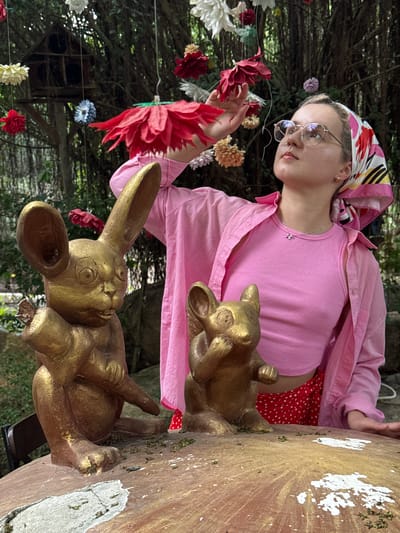 The image depicts a young woman in an outdoor, whimsical setting, likely in Nam Nha Trang, Vietnam. She is positioned on the left side of the frame, looking towards the right and slightly downwards, seemingly interacting with a vibrant red artificial flower that is strung up. Her expression is calm and contemplative.
The woman wears a colorful headscarf featuring patterns in pink, yellow, black, and white, and round, metal-rimmed glasses. She has a pink long-sleeved button-up shirt worn open over a pink ribbed tank top, and a silver necklace with a small, star-shaped pendant. A red polka-dot skirt is visible below her shirt.
To her right, two golden rabbit statues are prominently displayed on a wooden surface. The larger rabbit is in the foreground, and a smaller one is positioned behind it. This, along with numerous other artificial flowers in various colors (red, white, pink, blue, yellow, orange) that are strung overhead and in the foreground, creates a decorative and somewhat fantastical atmosphere, reminiscent of a garden party or themed attraction.
The background consists of blurred, lush green foliage, suggesting a garden or a densely planted outdoor area. The wooden surface where the rabbits rest shows signs of age with some moss and white paint splatters. The natural lighting indicates it is daytime. No visible text is present.