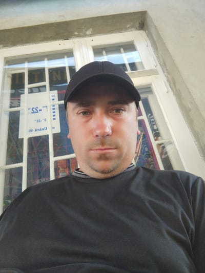 A man wearing a black baseball cap and a black long-sleeved shirt is shown in a close-up, waist-up shot. He has fair skin with some redness on his cheeks and nose, and a light stubble on his chin and upper lip. His hair is not visible due to the cap. His eyes are open and he is looking directly at the camera.
He is positioned in front of a large window with white mullions. Behind the window, there are visible bars, suggesting the window might be for a ground-floor establishment or a secured area. A white sign with blue text and graphics is partially visible on the window. The overall environment appears to be outdoors, possibly a street or an alley, with a concrete wall on the right side of the frame. The lighting suggests it is daytime. The location is identified as Costești, Moldova.