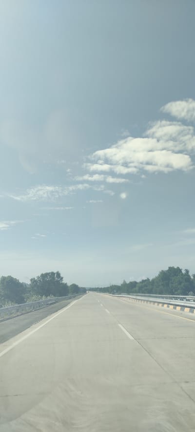 The image shows an empty highway stretching into the distance under a hazy blue sky with scattered white clouds. The highway is made of concrete and has white lane markings. On either side of the highway, there are guardrails and vegetation, consisting of trees and bushes. The scene suggests a daytime, possibly late morning or early afternoon, given the brightness and the haze. The environment appears rural or semi-rural. There are no people or vehicles visible in the frame, giving the impression of a peaceful, unpopulated road. There is a subtle lens flare in the upper right quadrant of the sky. The overall impression is one of openness and a long, straight path ahead. The location is specified as Gampongcot, Indonesia, suggesting this is a typical highway scene in that region.