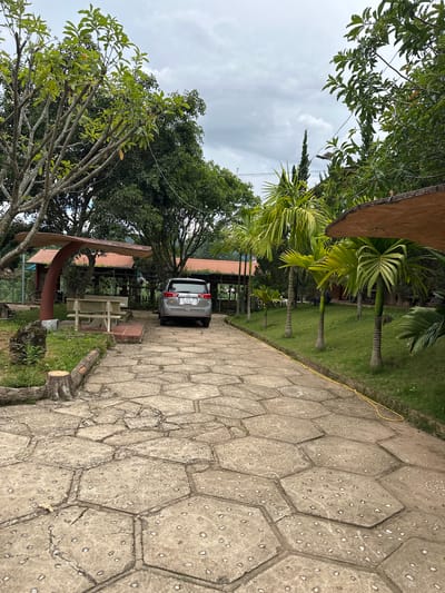 This image depicts a paved walkway leading towards a building complex in a rural setting. The walkway is constructed of hexagonal concrete slabs with small stones embedded in them, showing signs of cracking and wear. To the left of the walkway, there is a concrete picnic bench under a brown, mushroom-shaped canopy, and further back, a building with a red roof and open-air seating areas.
On the right side of the walkway, a grassy area is lined with several palm trees of varying heights. A silver SUV with license plate "51G 635.89" is parked on the paved area to the right of the building entrance. The background is filled with lush green trees and vegetation, suggesting a tropical or subtropical climate. The sky is overcast, with scattered clouds. The overall scene appears to be an outdoor seating or rest area, possibly part of a resort or restaurant, located in Thôn 2, Vietnam.