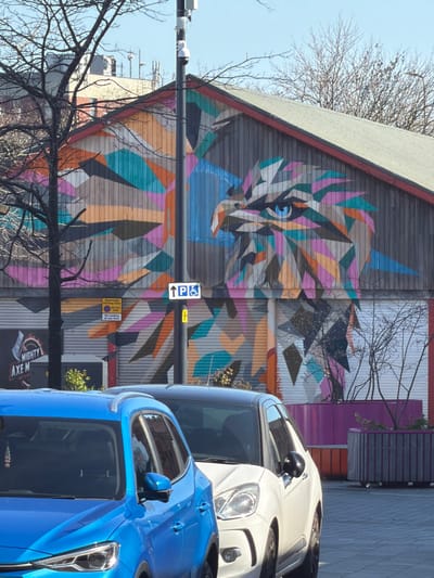 The image displays an outdoor urban scene in Crewe, United Kingdom, specifically depicting the side of a building identified as the former Crewe Market Hall, now part of the Crewe Lifestyle Centre. The building's facade is dominated by a large, geometric mural of an eagle's head in profile, facing right. The mural is composed of numerous irregular polygons in varied colors, including teal, orange, pink, purple, grey, black, and white, with the eagle's eye rendered in blue. Below the mural, sections of roll-down security shutters are visible, one featuring a partial sign reading "MIGHTY AXE M[E]".

In the foreground, two parked vehicles are partially visible: a blue SUV/crossover on the left and a white compact car on the right, both oriented away from the viewer. A tall, dark grey utility pole stands centrally, equipped with a CCTV camera and communication antennas. Attached to this pole is a white rectangular sign indicating disabled parking with an upward arrow. To the left of the pole, a "Restricted Parking Zone" sign is visible, stating "AT ALL TIMES / No loading at any time." A leafless tree with dark branches partially obscures the mural on the left. On the right, purple and grey wooden planters contain sparse, leafless shrubbery. The background shows a clear blue sky. No individuals are present in the scene.