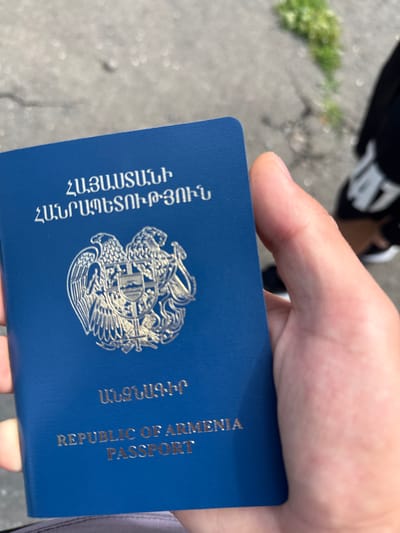 An Armenian passport is held in a person's hand against a blurred background of pavement and grass. The passport is dark blue with a silver embossed coat of arms in the center, and gold lettering above and below it. The text on the passport reads "ՀԱՅԱՍՏԱՆԻ ՀԱՆՐԱՊԵՏՈՒԹՅՈՒՆ" and "REPUBLIC OF ARMENIA PASSPORT". The person's thumb and index finger are visible on the left side of the image. The background appears to be an outdoor setting, possibly a street or walkway in the city of Tumanyan, Armenia. A person wearing dark clothing and sneakers is partially visible in the upper right corner of the image.