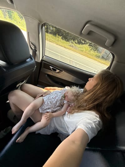 The image depicts the interior of a car, as evidenced by the black leather seats, door panel, and window. In the foreground, a woman with long, reddish-brown hair is seen from the side, lying down with her eyes closed. She is wearing a white t-shirt. Resting on her lap is a young child, who is also asleep, wearing a white dress with a pattern of small animals. The child's head is against the woman's chest, and their legs are extended. The background shows a view through the car window of trees and vegetation, suggesting a rural or suburban setting. The overall scene conveys a sense of peaceful rest during a car journey. The location in Khryastovo, Russia, is not visually apparent from the image itself.