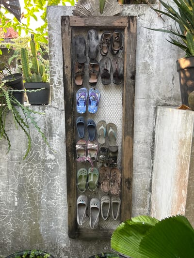 The image displays an outdoor scene in Banguntapan, Indonesia, featuring a rustic wooden frame with a wire mesh backing, mounted on a textured concrete wall. This frame serves as a makeshift shoe rack, displaying an assortment of footwear, primarily flip-flops and sandals, organized in vertical rows. The shoes vary in style, color, and condition, suggesting they are used. Surrounding the shoe rack, various plants, including cacti and leafy green foliage, are visible, some in pots and others growing wild. The background shows more concrete structures and hints of vegetation and rooftops, indicating an urban or semi-urban environment. The lighting suggests it is daytime, with sunlight casting shadows and highlighting textures.